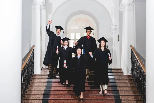 Alegre grupo de estudiantes en vestidos de graduación haciendo gestos mientras sostiene diplomas - foto de stock