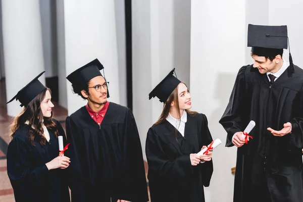 Homem alegre gesticulando perto de estudantes em vestidos de graduação e segurando diploma — Fotografia de Stock