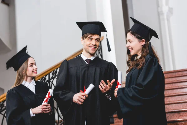 Fröhlicher Mann mit Abschlussmütze gestikuliert neben attraktiven Mädchen, während er sein Diplom in der Hand hält — Stockfoto