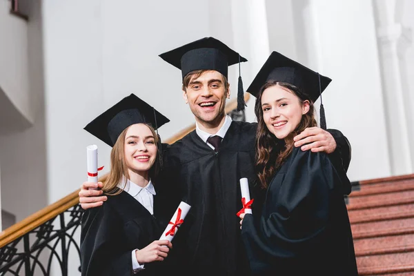Hombre alegre en gorra de graduación abrazando a chicas atractivas mientras sostiene el diploma - foto de stock