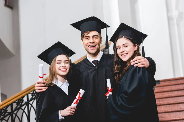 Homem alegre na formatura cap abraçando meninas atraentes enquanto segurando diploma — Fotografia de Stock