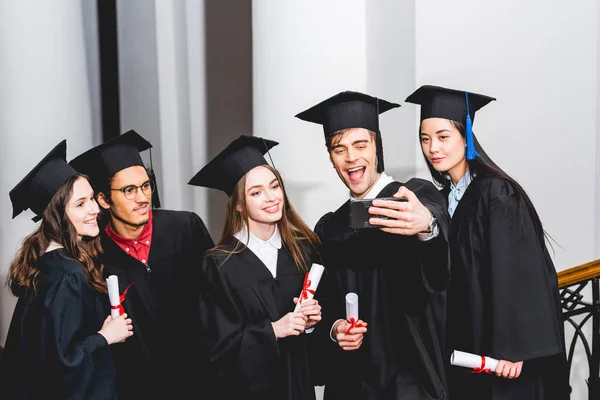 Cheerful students in graduation gowns taking selfie and smiling while holding diplomas — Stock Photo