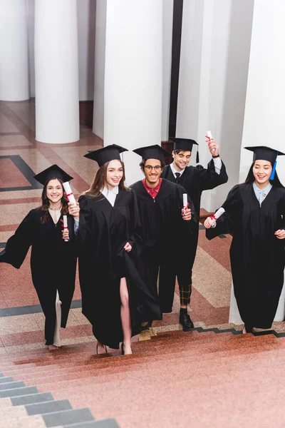 Cheerful students in graduation gowns holding diplomas and walking on stairs — Stock Photo