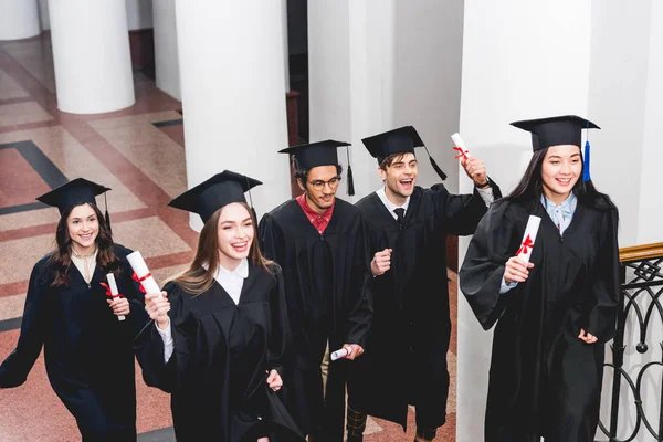Улыбающиеся студенты в выпускных платьях с дипломами в университете — стоковое фото