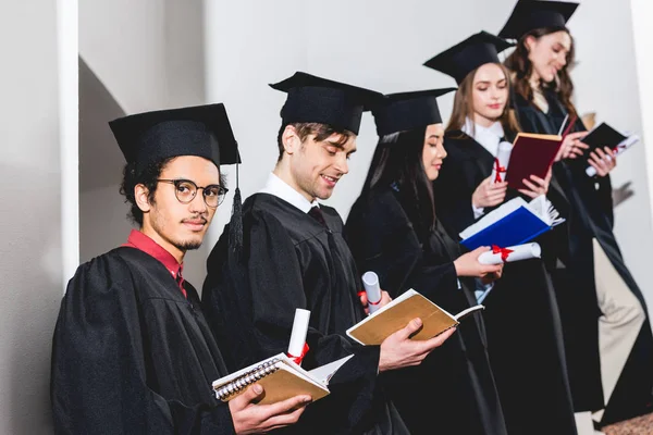 Vista de ángulo bajo del hombre en gafas mirando a la cámara cerca de los estudiantes en gorras de graduación lectura de libros - foto de stock