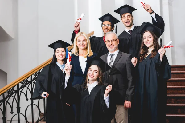 Studenti allegri in berretti di laurea titolari di diplomi vicino a insegnanti felici — Foto stock