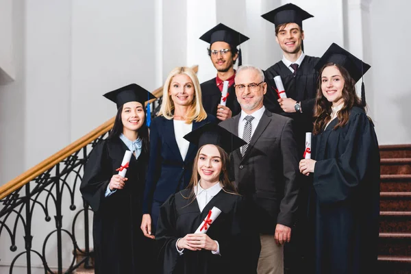 Усміхнені студенти у випускних шапках, що тримають дипломи біля щасливих вчителів — стокове фото