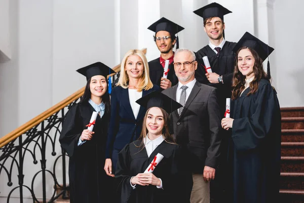 Étudiants souriants en casquettes de fin d'études détenant des diplômes près des enseignants heureux — Photo de stock