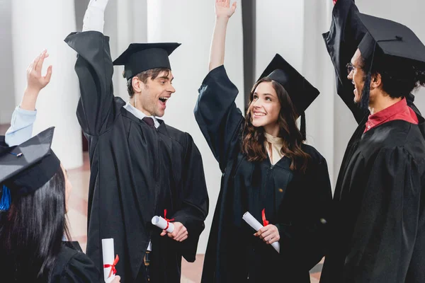 Grupo sonriente en los estudiantes en vestidos de graduación con diplomas y poner las manos por encima de la cabeza - foto de stock
