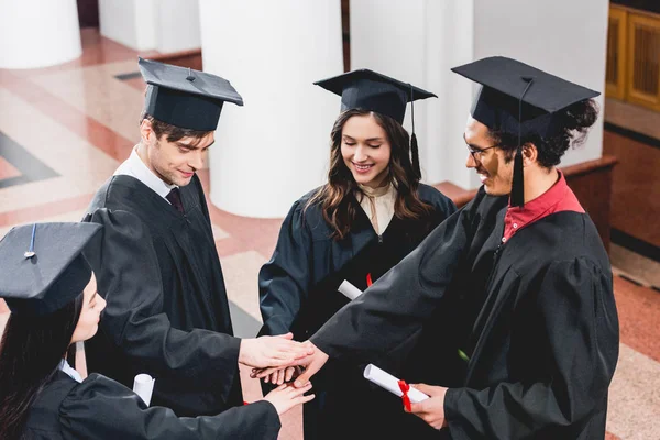 Vista aérea del grupo alegre en estudiantes que ponen las manos juntas mientras que sostienen diplomas - foto de stock