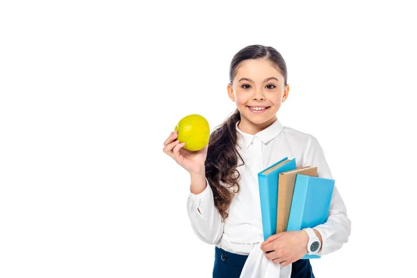 Lächelndes Schulmädchen mit Apfel und Büchern in der Hand, während es isoliert auf weiß mit Kopierraum in die Kamera blickt — Stockfoto