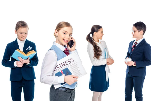 Colegiala con el periódico hablando en el teléfono inteligente cerca de los escolares que fingen ser empresarios aislados en blanco - foto de stock