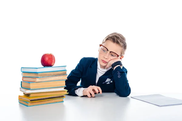 Triste écolier dans des lunettes assis au bureau avec des livres et pomme isolé sur blanc — Photo de stock