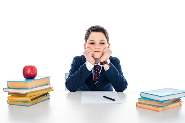 Sad schoolboy sitting at desk with books and propping chin isolated on white — Stock Photo