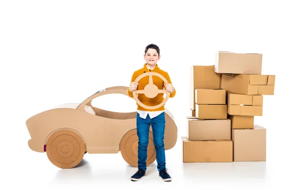 Boy near cardboard car holding steering wheel and looking at camera on white — Stock Photo