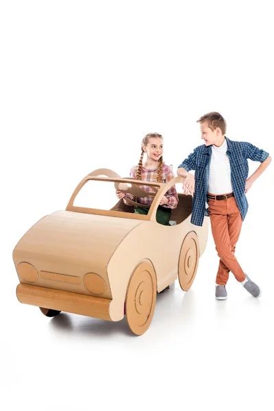 Smiling kid sitting in cardboard car and looking at boy on white with copy space — Stock Photo