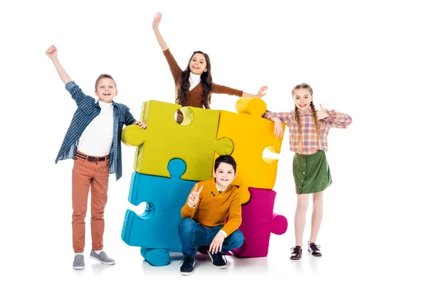 Crianças alegres posando perto de peças de quebra-cabeça coloridas no branco — Fotografia de Stock