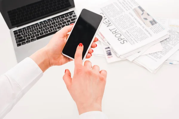 Enfoque selectivo de la mujer utilizando el teléfono inteligente con pantalla en blanco cerca de la computadora portátil y periódicos con noticias deportivas - foto de stock