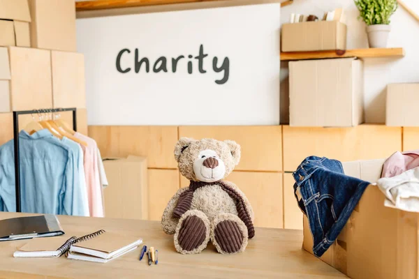 Caja de cartón con ropa, osito de peluche y cuadernos en mesa de madera cerca de pancarta con inscripción de caridad - foto de stock