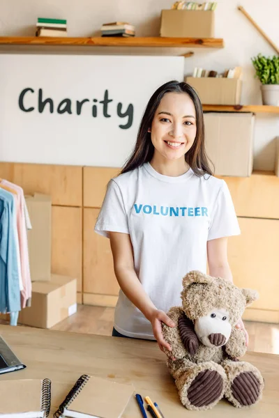 Pretty asian volunteer girl holding teddy bear, smiling and looking at camera — Stock Photo