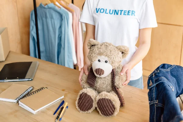 Vista recortada de una chica voluntaria sosteniendo un oso de peluche mientras está de pie cerca de una mesa de madera con cuadernos - foto de stock