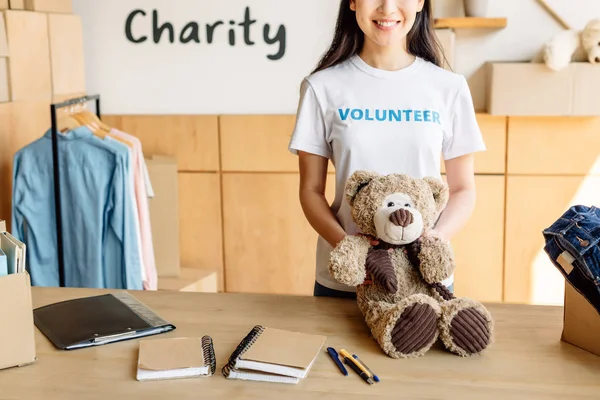Young volunteer girl standing with teddy bear near wooden table with notebooks — Stock Photo