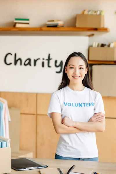 Attractive asian girl in white t-shirt with volunteer inscription standing with crossed arms and looking at camera — Stock Photo