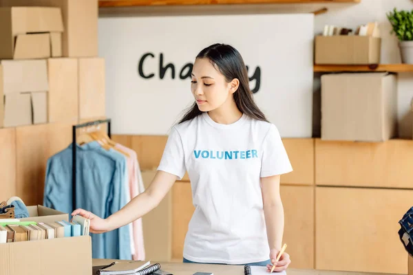 Attentive asian volunteer writing in notebook while standing near rack with shirts — Stock Photo