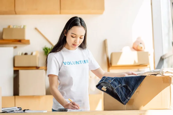 Concentrated asian volunteer writing in notebook while standing near carton box with clothes — Stock Photo