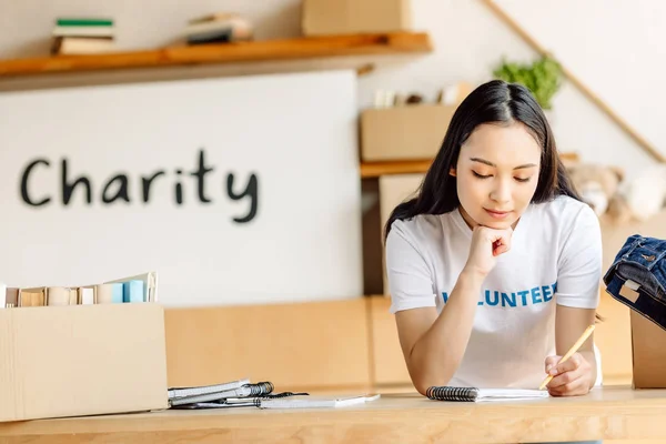Thoughtful asian volunteer writing in notebook while standing near carton boxes with clothes and books — Stock Photo