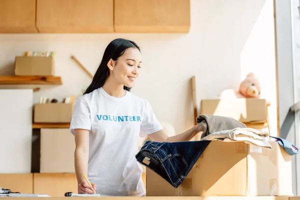 Lächelnder asiatischer Freiwilliger schreibt in Notizbuch, während er neben Karton mit Kleidung steht — Stockfoto