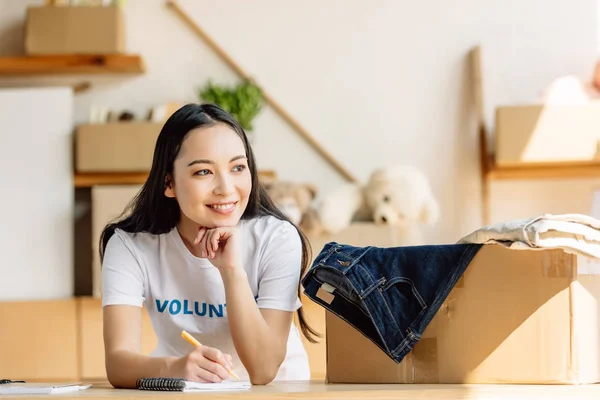 Beautiful asian volunteer writing in notebook and lookin away while standing near carton box with clothes — Stock Photo