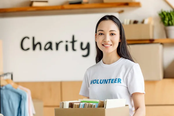 Beautiful asian volunteer holding cardboard box with books and looking at camera — Stock Photo