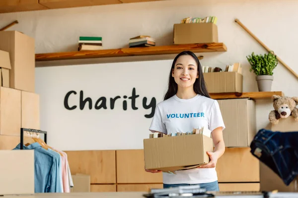 Smiling asian volunteer holding cardboard box with books and looking at camera — Stock Photo
