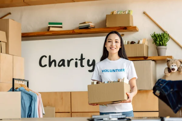 Sonriente asiático voluntario celebración de cartón caja con libros y mirando a cámara - foto de stock