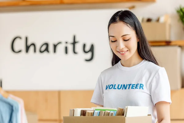 Attractive asian volunteer holding cardboard box with books and looking down — Stock Photo