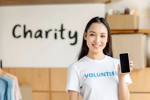 Beau bénévole asiatique tenant smartphone avec écran blanc, souriant et regardant la caméra — Photo de stock