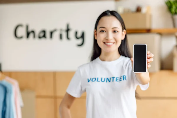 Enfoque selectivo de bastante asiático voluntario sosteniendo teléfono inteligente con pantalla en blanco, sonriendo y mirando a la cámara - foto de stock
