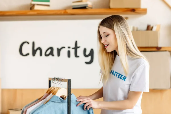 Pretty blonde volunteer standing near rack with different shirts — Stock Photo