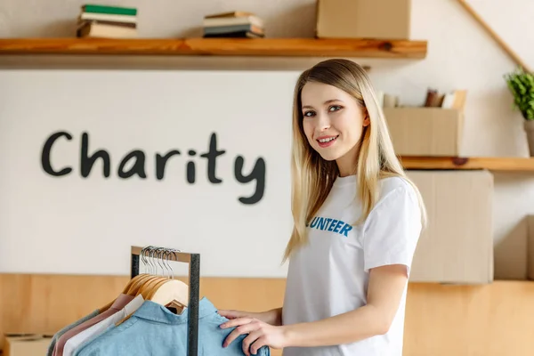 Beautiful blonde volunteer standing near rack with shirts and looking at camera — Stock Photo