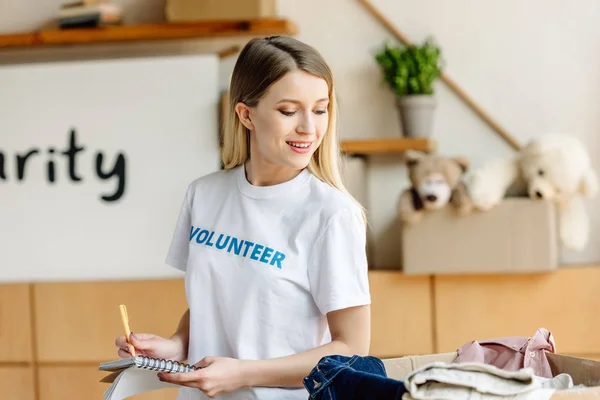 Pretty volunteer writing in notebook while standing near carton box with clothes — Stock Photo