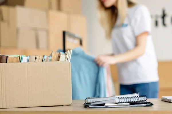 Selective focus of cardboard box with books, and volunteer girl standing near rack with clothes — Stock Photo