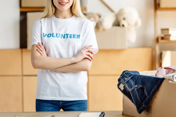 Cropped view of volunteer standing with crossed arms near carton box with clothes — Stock Photo