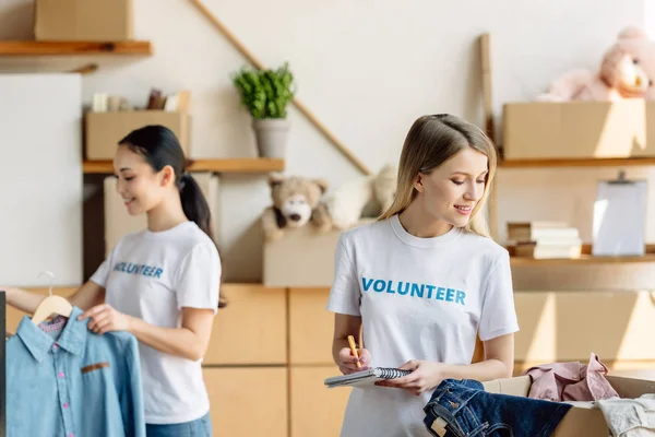 Selective focus of beautiful young volunteer writing in notebook near asian girl standing by rack with clothes — Stock Photo