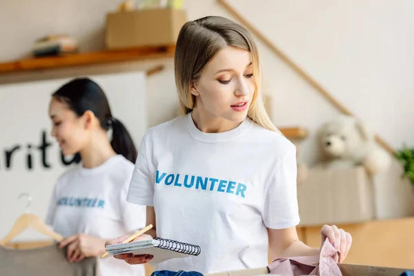 Selective focus of attractive young volunteer writing in notebook near asian girl standing by rack with clothes — Stock Photo