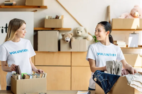 Zwei schöne multikulturelle Freiwillige beim Auspacken von Kartons mit Kleidung und Büchern — Stockfoto