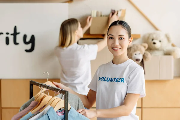 Selective focus of pretty asian volunteer standing near rack with shirts and looking at camera — Stock Photo