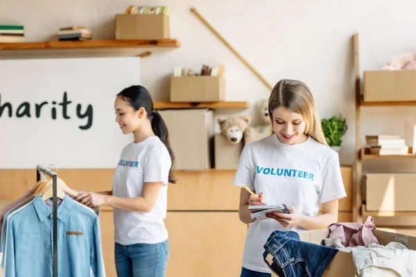 Selective focus of beautiful young volunteer writing in notebook near asian girl standing by rack with clothes — Stock Photo