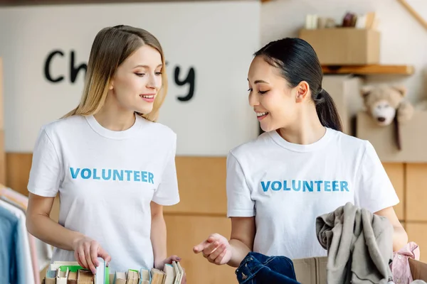 Two attractive multicultural volunteers unpacking carton boxes with books and clothes — Stock Photo