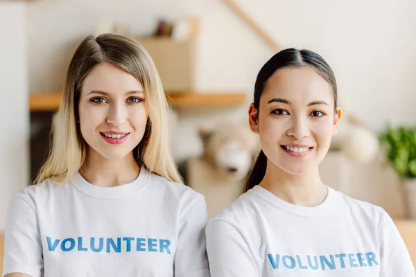 Deux beaux bénévoles multiculturels en t-shirts blancs souriants et regardant la caméra — Photo de stock